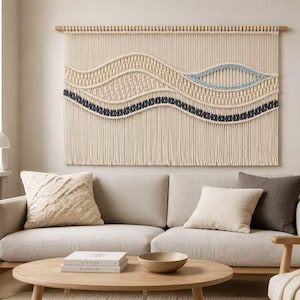 May include: A large rectangular macrame wall hanging suspended from a wooden dowel. The piece features off-white cotton cord with intricate knot patterns, including a prominent wave design accented by a dark charcoal gray line and a light blue oval.