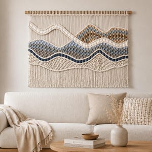 Puede incluir: Un tapiz de macramé rectangular de gran tamaño con un diseño de paisaje que presenta capas onduladas de cordón anudado en color crema, azul claro, azul oscuro, tostado y marrón. La pieza cuelga de un listón de madera y tiene flecos.