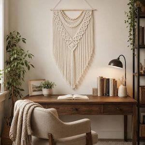 May include: A large off-white macrame wall hanging featuring intricate knotted patterns, a row of wooden beads, and long fringe. The piece is suspended from a horizontal wooden dowel with a string, creating a V-shaped design on the wall.