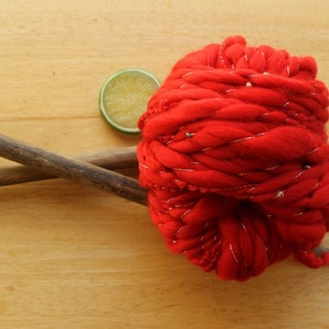 A skein of handspun, solid red, thick and thin yarn. The super bulky yarn is plied with silver thread and glass beads. It is resting on a light wood background with a pair of large, wooden knitting needles and a lime slice.