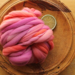 A skein of handspun, super bulky, thick and thin yarn. The yarn is hand dyed in self striping lilac and coral. The yarn is resting on a handmade, wooden plate, on a light wood background with a lime slice.