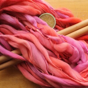A hank of handspun, super bulky, thick and thin yarn. The yarn is hand dyed in self striping lilac and coral. The yarn is resting on a light wood background with a pair of large, wooden knitting needles and a lime slice.