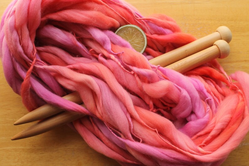 A hank of handspun, super bulky, thick and thin yarn. The yarn is hand dyed in self striping lilac and coral. The yarn is resting on a light wood background with a pair of large, wooden knitting needles and a lime slice.
