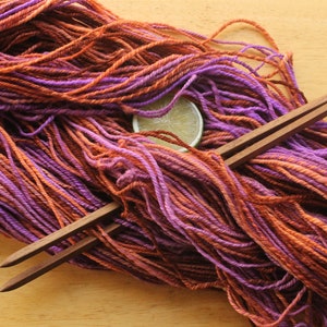 A hank of worsted weight, 2 ply yarn in lavender, peach, and rust. The yarn is laid diagonally across a light wood background with a pair of square, wooden knitting needles and a lime slice.