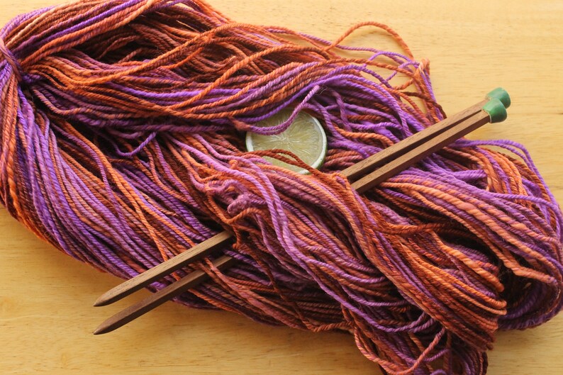 A hank of worsted weight, 2 ply yarn in lavender, peach, and rust. The yarn is laid diagonally across a light wood background with a pair of square, wooden knitting needles and a lime slice.