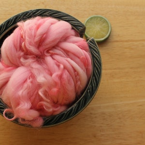 A skein of super bulky, hand dyed, handspun, thick and thin yarn. The yarn is bubblegum pink with wool curls. It is nestled in a dark green, ceramic yarn bowl on a light wood background with a lime slice.