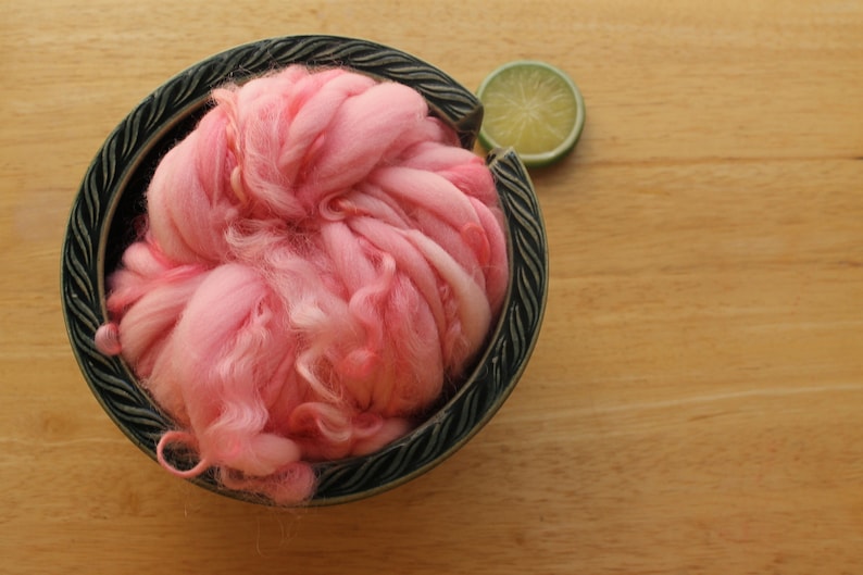 A skein of super bulky, hand dyed, handspun, thick and thin yarn. The yarn is bubblegum pink with wool curls. It is nestled in a dark green, ceramic yarn bowl on a light wood background with a lime slice.