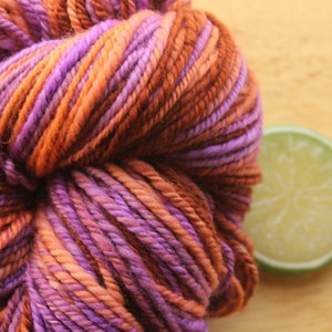 An extreme close up of a skein of worsted weight, 2 ply yarn in lavender, peach, and rust. The yarn is on a light wood background with a lime slice.