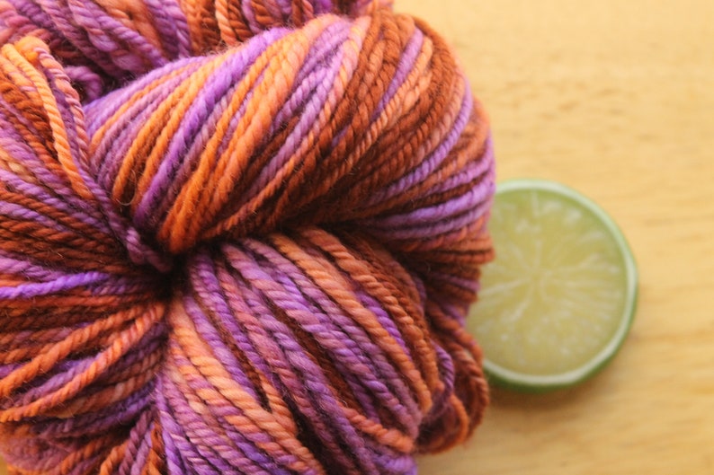 An extreme close up of a skein of worsted weight, 2 ply yarn in lavender, peach, and rust. The yarn is on a light wood background with a lime slice.