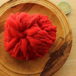 A skein of handspun, solid red, thick and thin yarn. The super bulky yarn is plied with silver thread and glass beads. It is resting on a handmade, wooden plate on a light wood background with a lime slice.