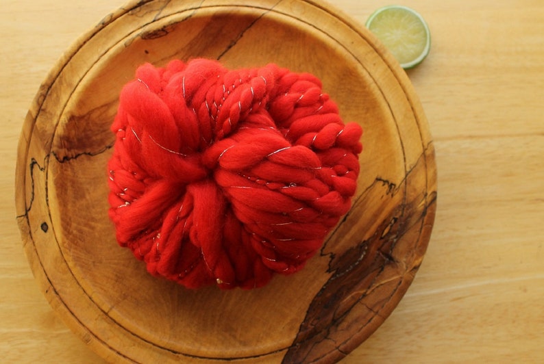 A skein of handspun, solid red, thick and thin yarn. The super bulky yarn is plied with silver thread and glass beads. It is resting on a handmade, wooden plate on a light wood background with a lime slice.