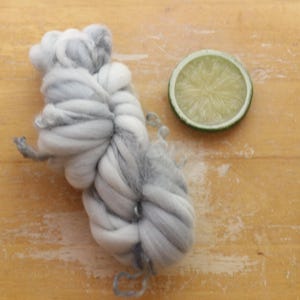 May include: A skein of gray and white yarn is displayed next to a lime slice on a wooden surface. The yarn is thick and twisted, with varying shades of gray and white. The lime slice is green and circular.