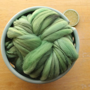 A skein of thick and thin, super bulky, handspun yarn. The yarn is hand dyed in dusty greens. It is nestled in a pale blue, ceramic yarn bowl on a light wood background with a lime slice.