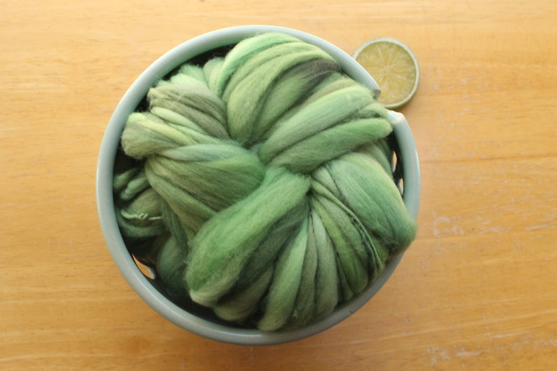 A skein of thick and thin, super bulky, handspun yarn. The yarn is hand dyed in dusty greens. It is nestled in a pale blue, ceramic yarn bowl on a light wood background with a lime slice.