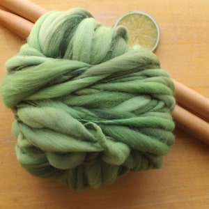 A skein of thick and thin, super bulky, handspun yarn. The yarn is hand dyed in dusty greens. It is resting on a light wood background with a pair of large, wooden knitting needles and a lime slice.