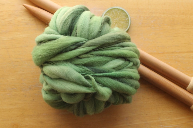 A skein of thick and thin, super bulky, handspun yarn. The yarn is hand dyed in dusty greens. It is resting on a light wood background with a pair of large, wooden knitting needles and a lime slice.