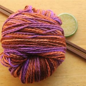 A skein of worsted weight, 2 ply yarn in lavender, peach, and rust. The yarn is resting on a light wood background with a pair of square, wooden knitting needles and a lime slice.