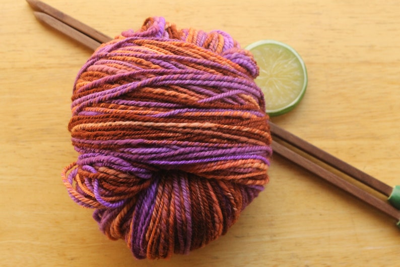 A skein of worsted weight, 2 ply yarn in lavender, peach, and rust. The yarn is resting on a light wood background with a pair of square, wooden knitting needles and a lime slice.