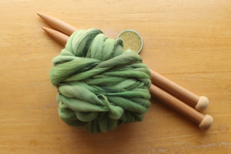 A skein of thick and thin, super bulky, handspun yarn. The yarn is hand dyed in dusty greens. It is resting on a light wood background with a pair of large, wooden knitting needles and a lime slice.