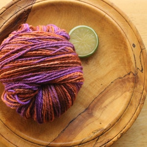 A skein of worsted weight, 2 ply yarn in lavender, peach, and rust. The yarn is resting on a wooden plate on a light wood background with a lime slice.