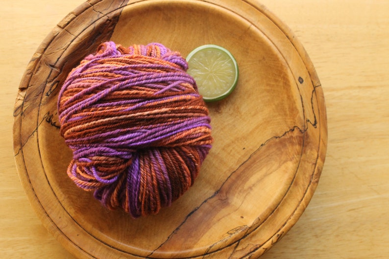 A skein of worsted weight, 2 ply yarn in lavender, peach, and rust. The yarn is resting on a wooden plate on a light wood background with a lime slice.