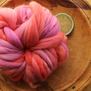 A skein of handspun, super bulky, thick and thin yarn. The yarn is hand dyed in self striping lilac and coral. The yarn is resting on a handmade, wooden plate, on a light wood background with a lime slice.