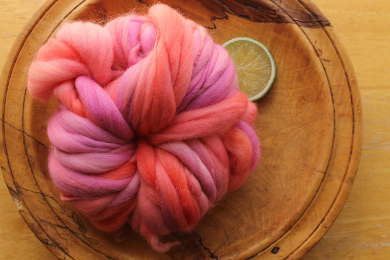 A skein of handspun, super bulky, thick and thin yarn. The yarn is hand dyed in self striping lilac and coral. The yarn is resting on a handmade, wooden plate, on a light wood background with a lime slice.