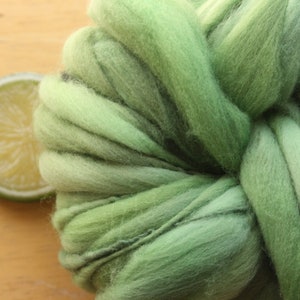 An extreme close up of a skein of thick and thin, super bulky, handspun yarn. The yarn is hand dyed in dusty greens. It is resting on a light wood background with a lime slice.