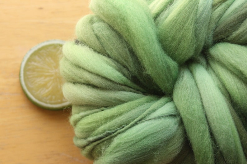 An extreme close up of a skein of thick and thin, super bulky, handspun yarn. The yarn is hand dyed in dusty greens. It is resting on a light wood background with a lime slice.