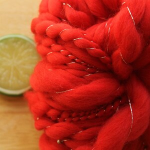 An extreme close up of a skein of handspun, solid red, thick and thin yarn. The super bulky yarn is plied with silver thread and glass beads. It is resting on a light wood background with a lime slice.