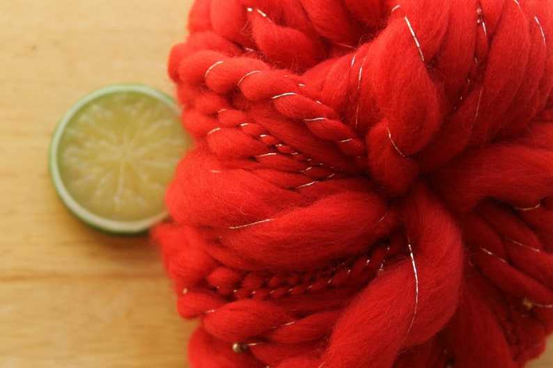 An extreme close up of a skein of handspun, solid red, thick and thin yarn. The super bulky yarn is plied with silver thread and glass beads. It is resting on a light wood background with a lime slice.