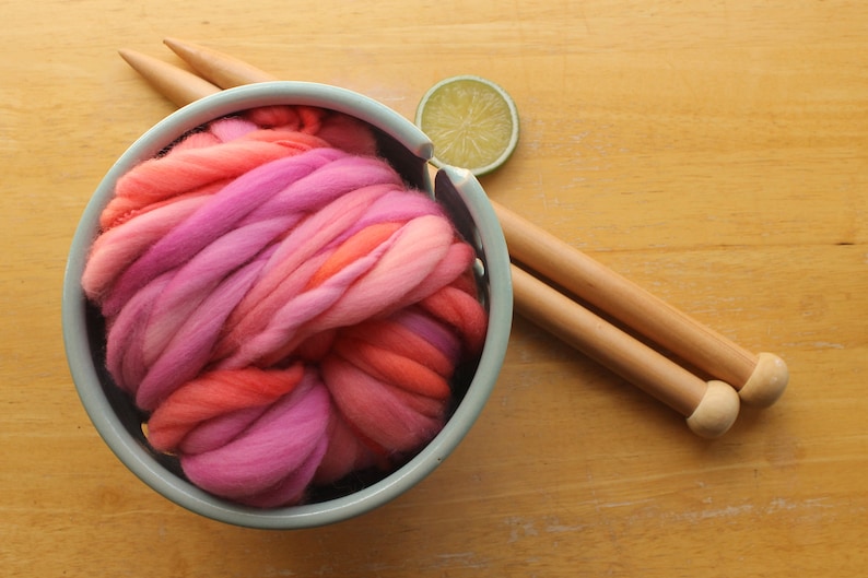 A skein of handspun, super bulky, thick and thin yarn. The yarn is hand dyed in self striping lilac and coral. The yarn is nestled in a ceramic yarn bowl on a light wood background with a pair of large, wooden knitting needles and a lime slice.