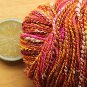 An extreme close up of a skein of handspun, worsted weight, 2 ply yarn in red, hot pink, yellow, and white.  The yarn is resting on a light wood background with a lime slice.