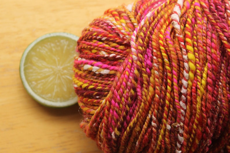 An extreme close up of a skein of handspun, worsted weight, 2 ply yarn in red, hot pink, yellow, and white.  The yarn is resting on a light wood background with a lime slice.