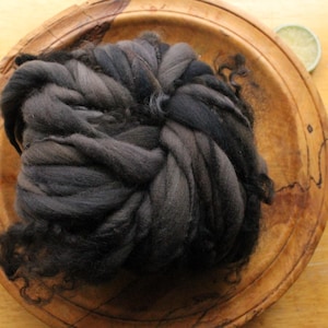 May include: A dark gray wool roving sits in a wooden bowl. The wool is tightly coiled, showing variations in color from charcoal to lighter shades of gray. The bowl is made of light brown wood.