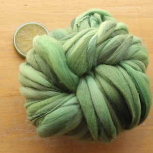 A skein of thick and thin, super bulky, handspun yarn. The yarn is hand dyed in dusty greens. It is resting on a light wood background with a lime slice.