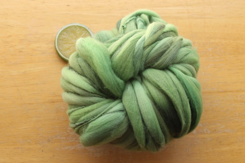 A skein of thick and thin, super bulky, handspun yarn. The yarn is hand dyed in dusty greens. It is resting on a light wood background with a lime slice.