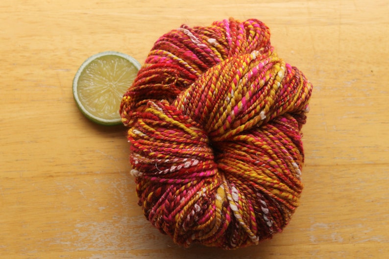 A skein of handspun, worsted weight, 2 ply yarn in red, hot pink, yellow, and white.  The yarn is resting on a light wood background with a lime slice.