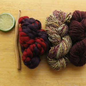 May include: Three skeins of yarn in different colors and textures. One skein is a deep red and purple, another is a variegated green, yellow, and red, and the third is a solid dark red.