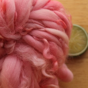 An extreme close up of a skein of super bulky, hand dyed, handspun, thick and thin yarn. The yarn is bubblegum pink with wool curls. It is laying on a light wood background with a lime slice.