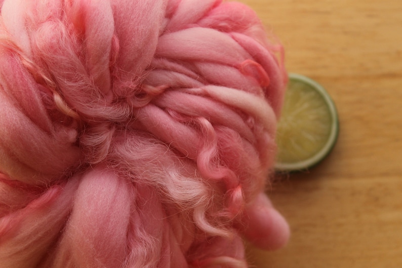 An extreme close up of a skein of super bulky, hand dyed, handspun, thick and thin yarn. The yarn is bubblegum pink with wool curls. It is laying on a light wood background with a lime slice.