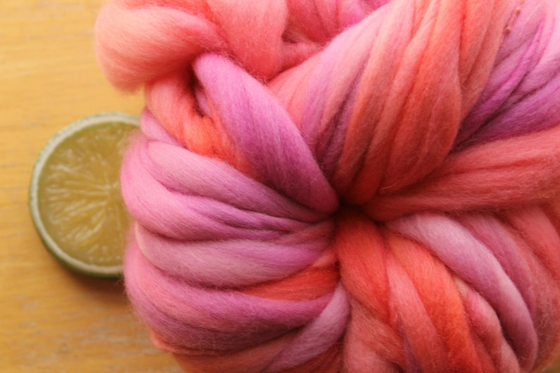 An extreme close up of a skein of handspun, super bulky, thick and thin yarn. The yarn is hand dyed in self striping lilac and coral. The yarn is resting on a light wood background with a lime slice.