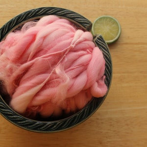 A skein of super bulky, hand dyed, handspun, thick and thin yarn. The yarn is bubblegum pink with wool curls. It is nestled in a dark green, ceramic yarn bowl on a light wood background with a lime slice.