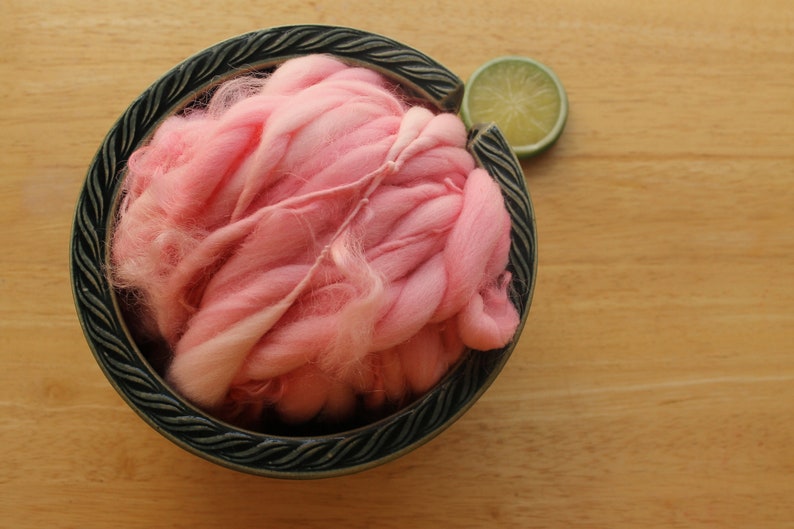 A skein of super bulky, hand dyed, handspun, thick and thin yarn. The yarn is bubblegum pink with wool curls. It is nestled in a dark green, ceramic yarn bowl on a light wood background with a lime slice.