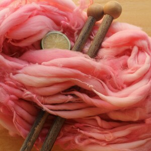 A hank of super bulky, hand dyed, handspun, thick and thin yarn. The yarn is bubblegum pink with wool curls. It is laying diagonally across a light wood background with a pair of large, wooden knitting needles and a lime slice.