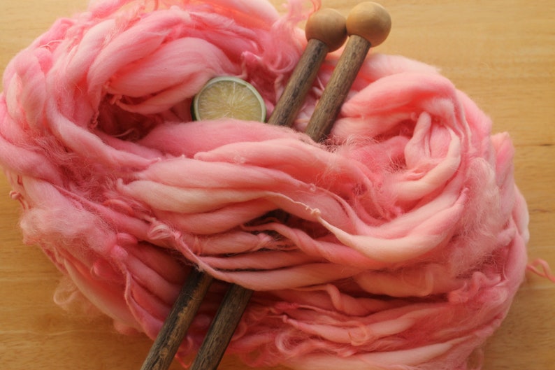 A hank of super bulky, hand dyed, handspun, thick and thin yarn. The yarn is bubblegum pink with wool curls. It is laying diagonally across a light wood background with a pair of large, wooden knitting needles and a lime slice.