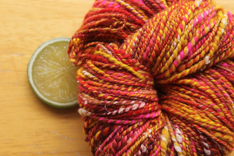 An extreme close up of a skein of handspun, worsted weight, 2 ply yarn in red, hot pink, yellow, and white.  The yarn is resting on a light wood background with a lime slice.