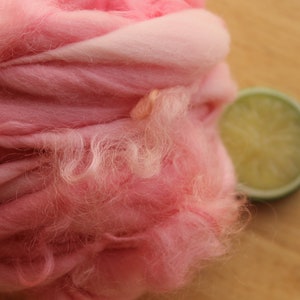 An extreme close up of a skein of super bulky, hand dyed, handspun, thick and thin yarn. The yarn is bubblegum pink with wool curls. It is laying on a light wood background with a lime slice.