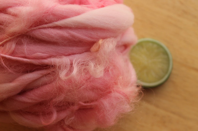 An extreme close up of a skein of super bulky, hand dyed, handspun, thick and thin yarn. The yarn is bubblegum pink with wool curls. It is laying on a light wood background with a lime slice.