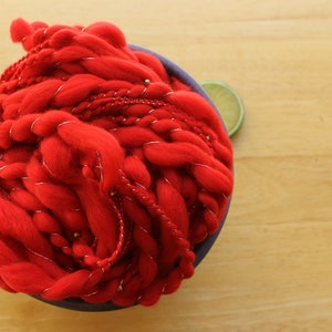 A hank of handspun, solid red, thick and thin yarn. The super bulky yarn is plied with silver thread and glass beads. It is nestled in a blue, ceramic yarn bowl on a light wood background with a lime slice.